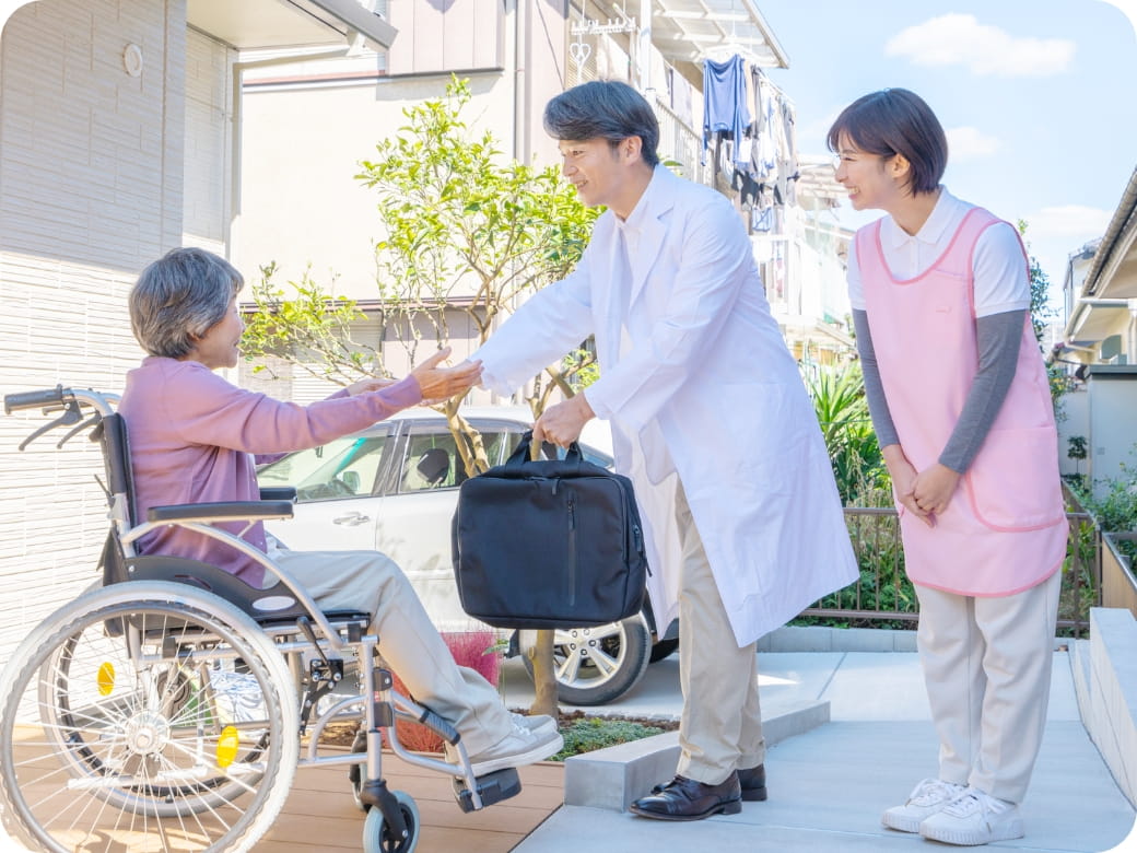 車椅子の高齢女性に向けて、医師が挨拶し、介護士が隣でサポートしている屋外シーン