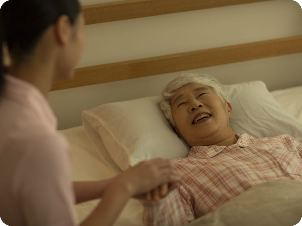 ベッドに横たわる高齢女性が笑顔で介護士に感謝の気持ちを伝えている場面