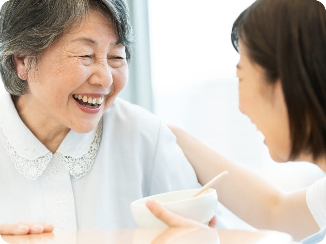 介護士がスプーンで食事をサポートしながら、高齢女性が笑顔で楽しそうにしている瞬間
