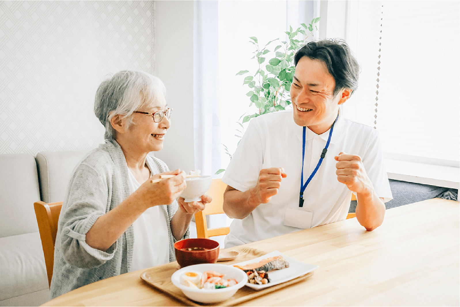 介護士と高齢女性がダイニングテーブルで笑顔で食事を楽しんでいる様子。