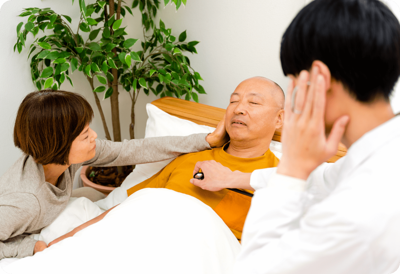 医師がベッドに横たわる男性の診察をしている様子。付き添いの女性が男性の手を優しく握り、サポートしている姿